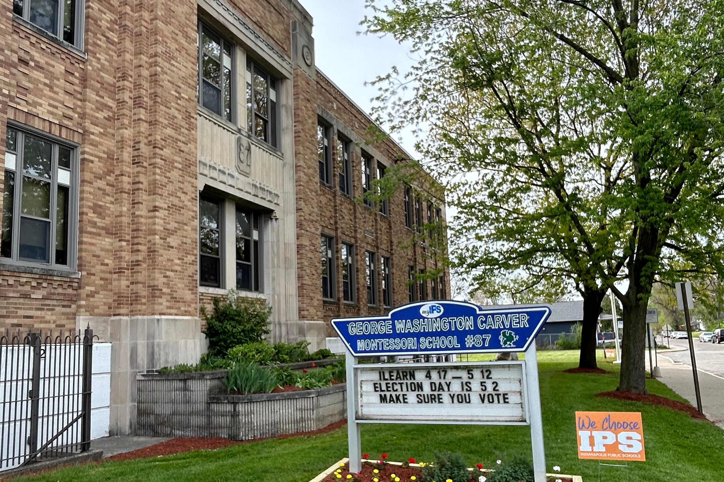 A blue and white sign in a green lawn in front of a large stone school building.