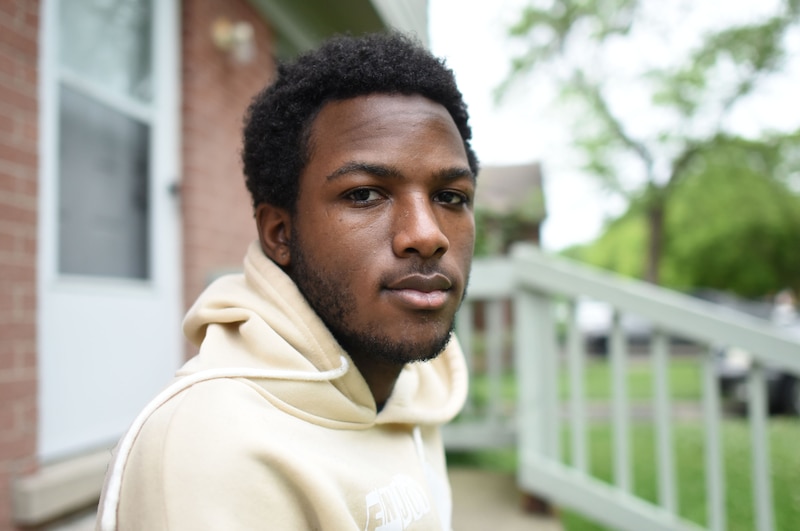 A young person with short dark hair and wearing a tan hoodie, poses for a portrait outside.