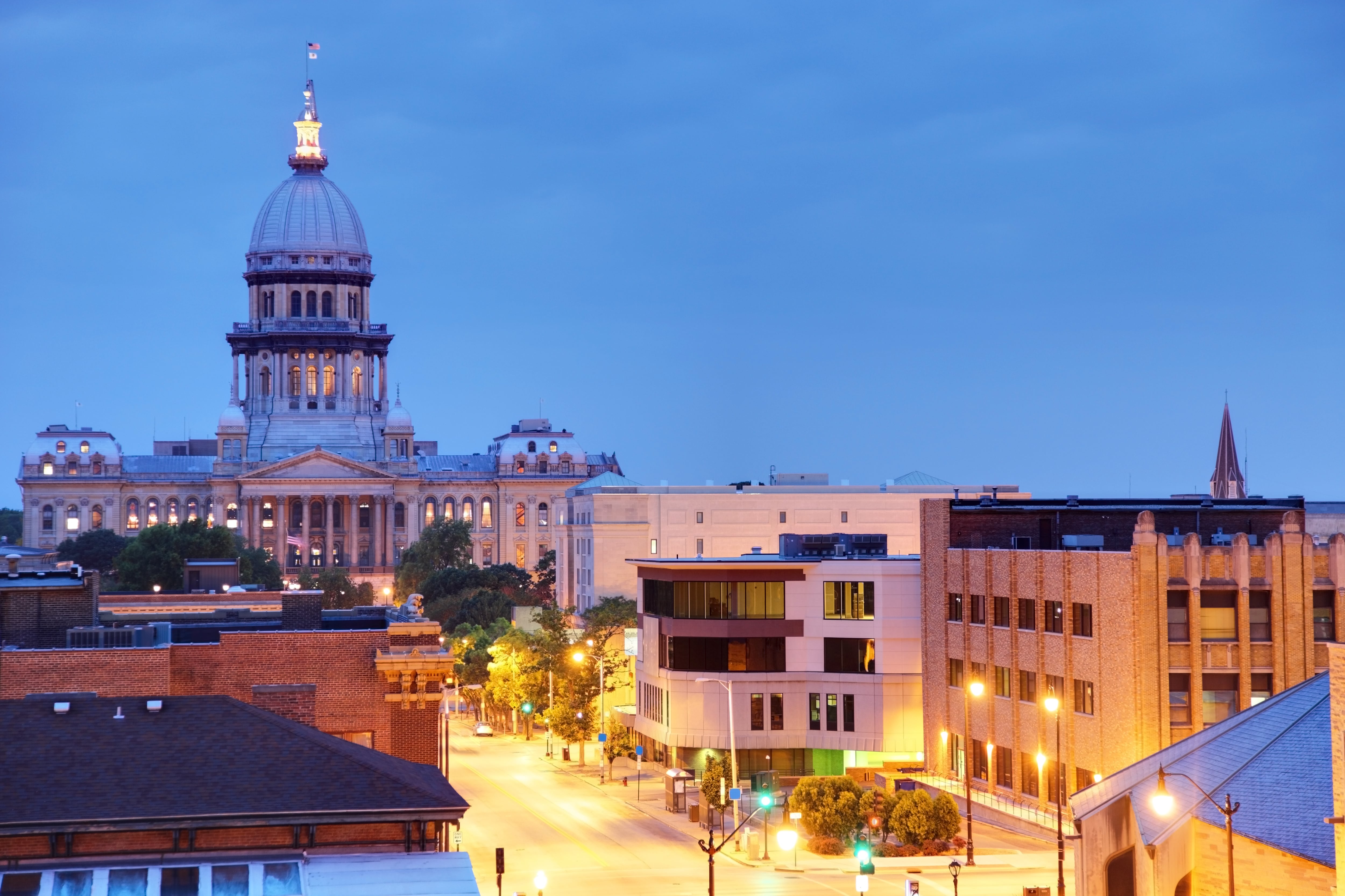 The Illinois State Capitol is seen along with other buildings and a street illuminated during the early evening.