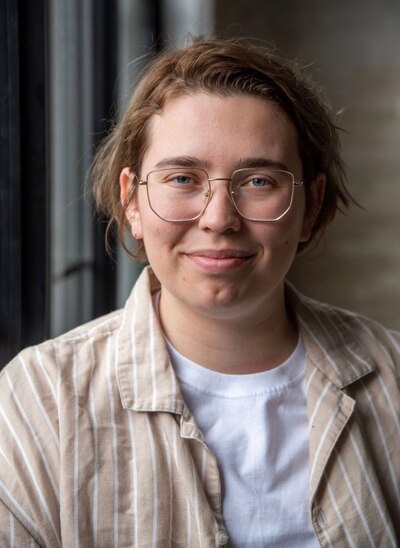 Photograph of a non-binary person wearing a white shirt and a tan and white striped button-down. They have light brown hair and wear glasses.