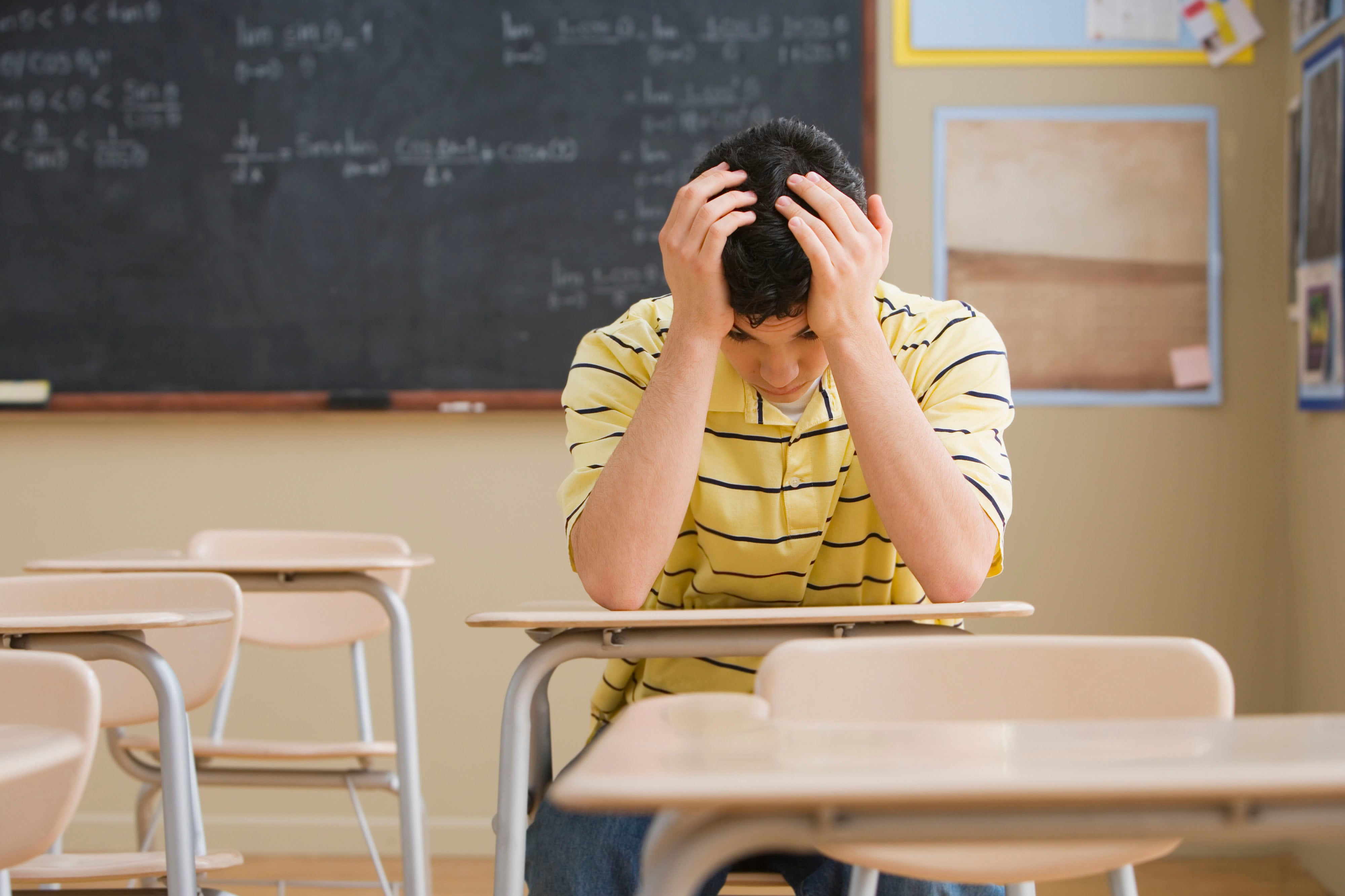 A teen boy wearing a yellow shirt holds his head in his hands while sitting at a wooden desk in a classroom.