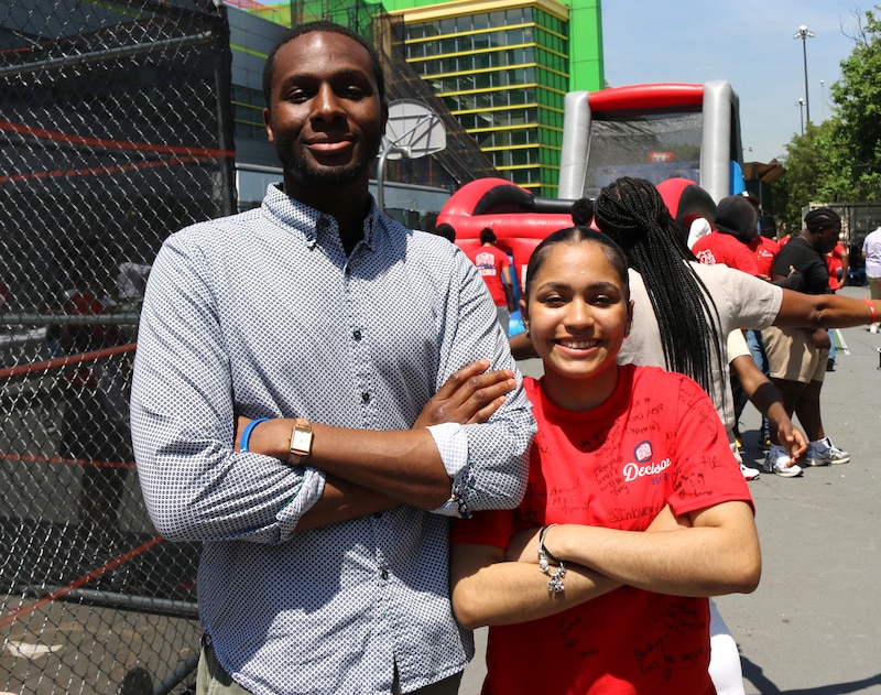 A man in a button down shirt and a young woman in a red shirt stand with their arms folded.