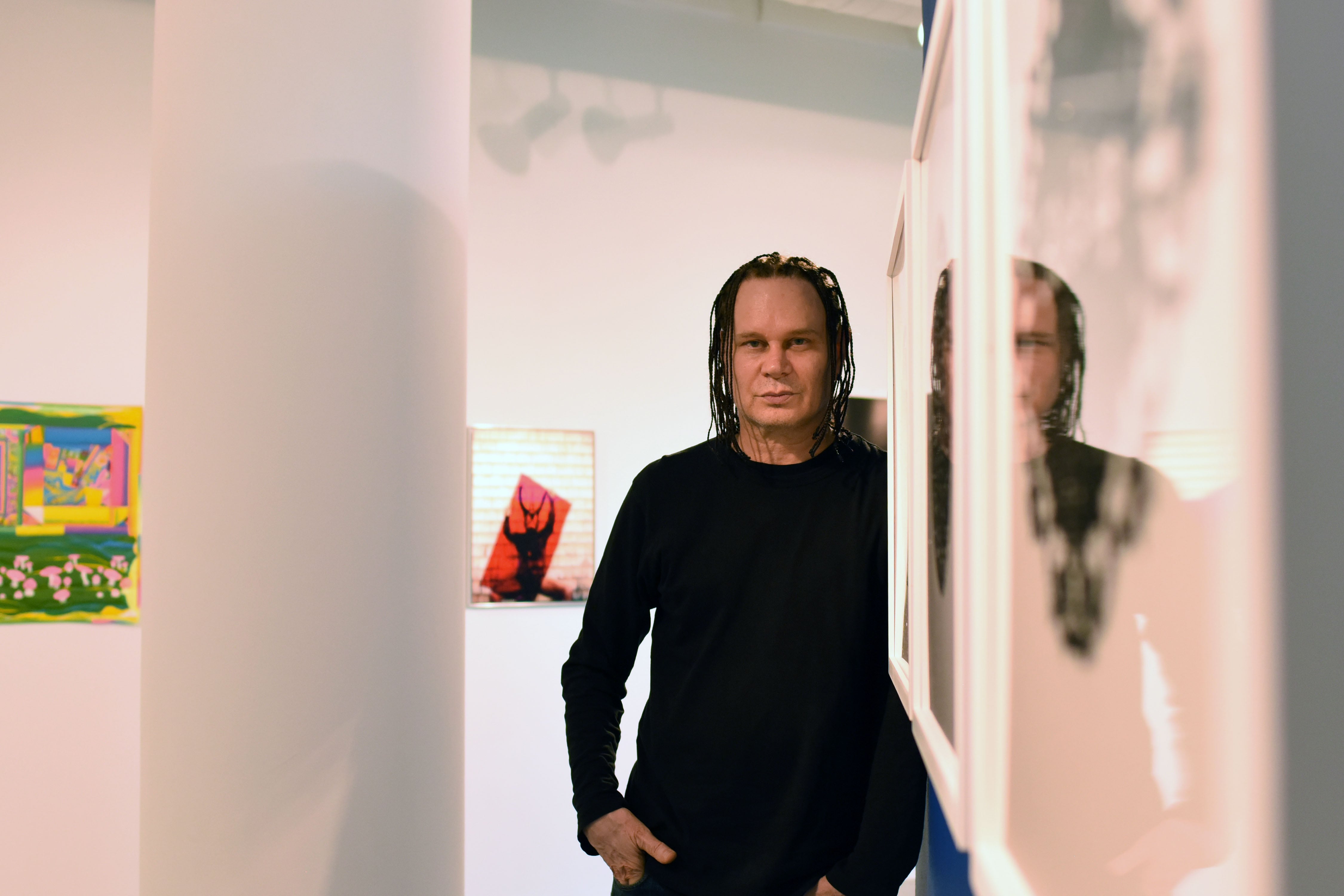 A person wearing all black poses for a portrait leaning up against an art gallery wall with art on the walls in the background.