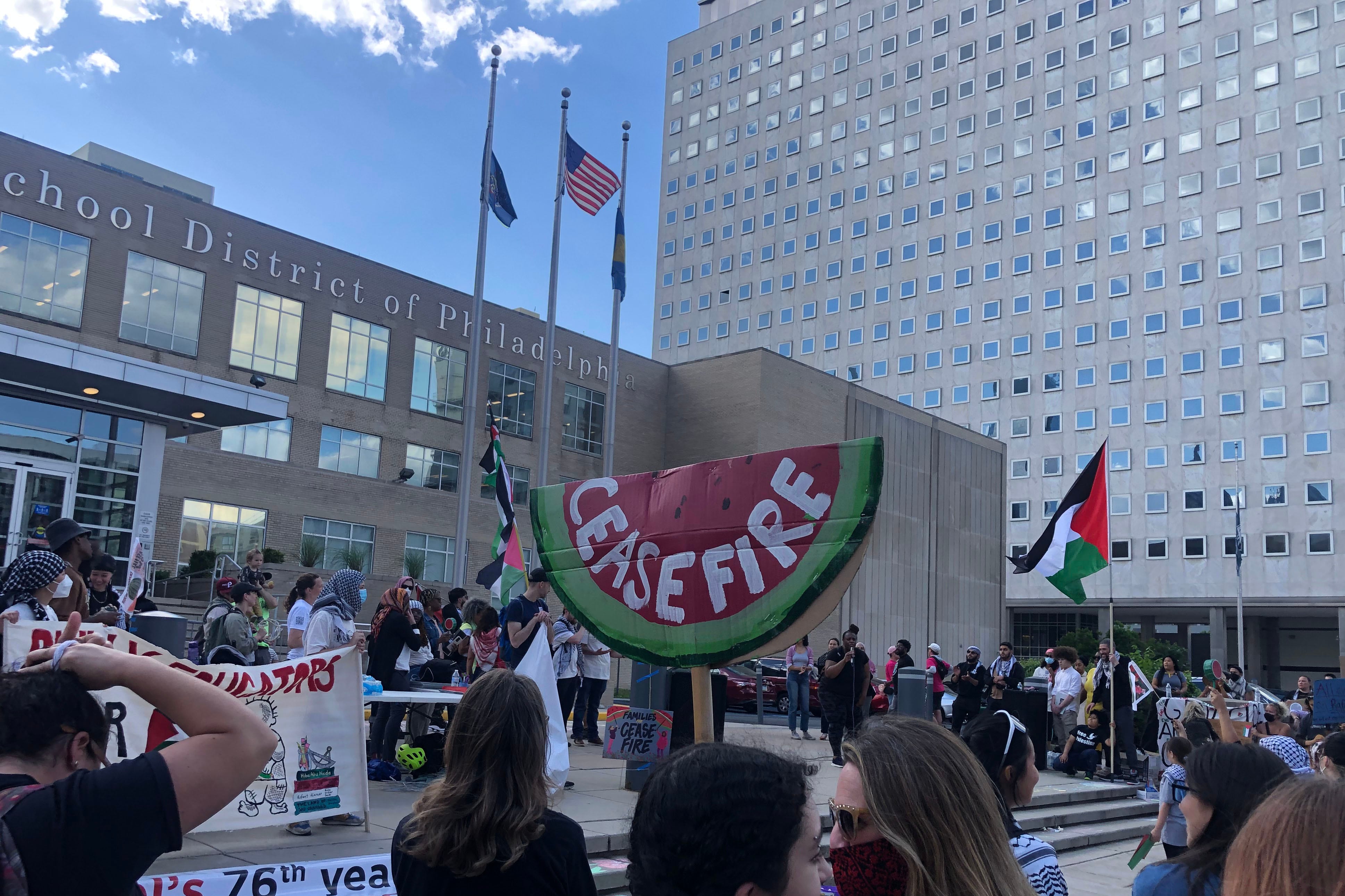 A large group of people protest outside a large stone building. Some people are wearing keffiyehs, holding signs and Palestinian flags.