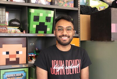 Un homme aux cheveux noirs courts, portant des lunettes et une chemise noire est assis devant une étagère avec des souvenirs de Minecraft.
