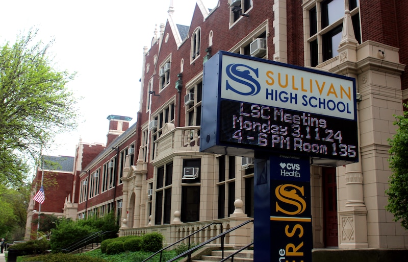 A white sign with blue and yellow letters stands outside a large red brick school building.