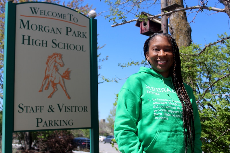 A woman with long dark hair and wearing a bright green hoodie poses for a portrait in front of a large white school sign outside.