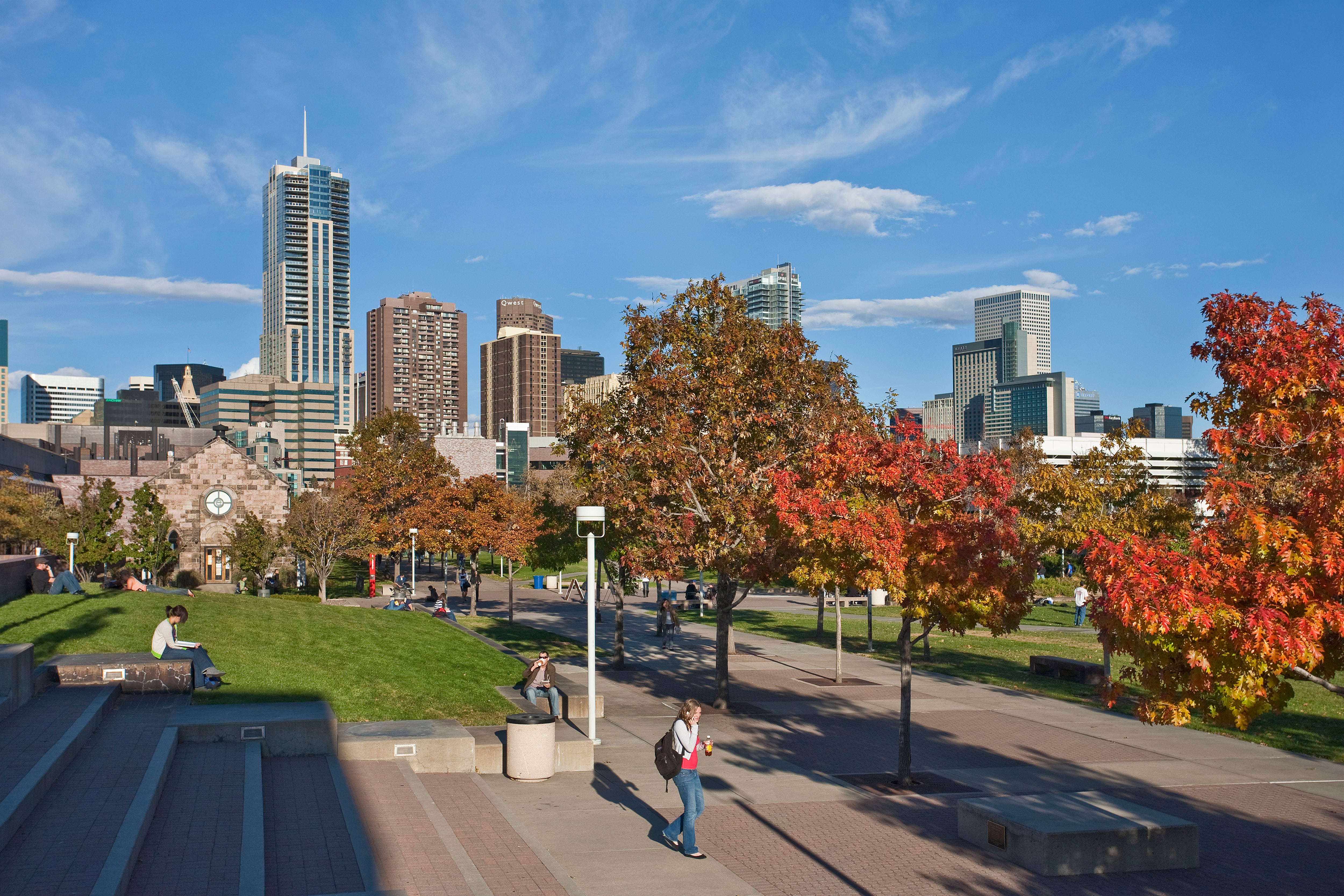 A college student wearing a backpack walks along a sidewalk with trees and Denver's skyline in the background.