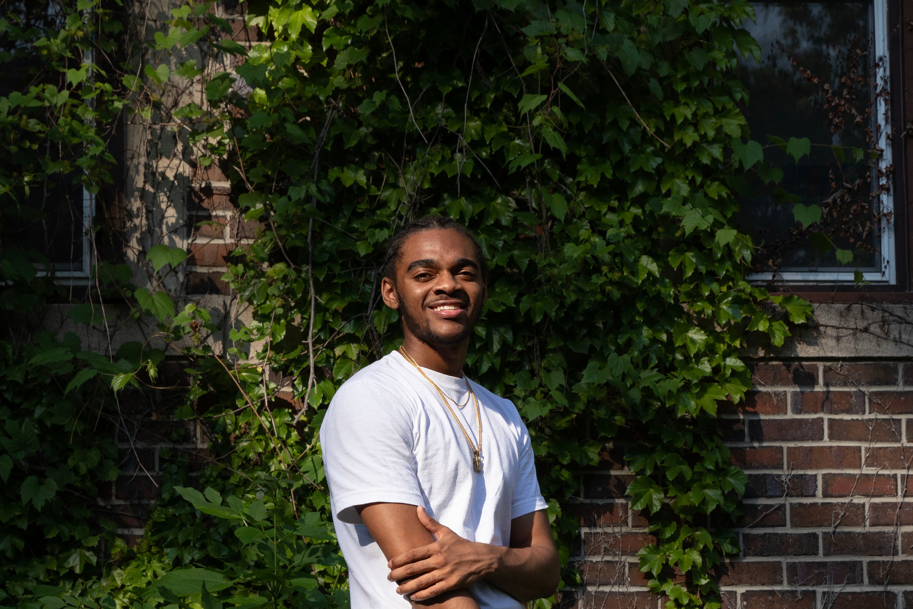 A high school senior with short dark hair and wearing a white t-shirt and a gold necklace with a diamond letter "K" crosses his arms and poses for a portrait while standing in front of a brick wall with greenery crawling up the wall.