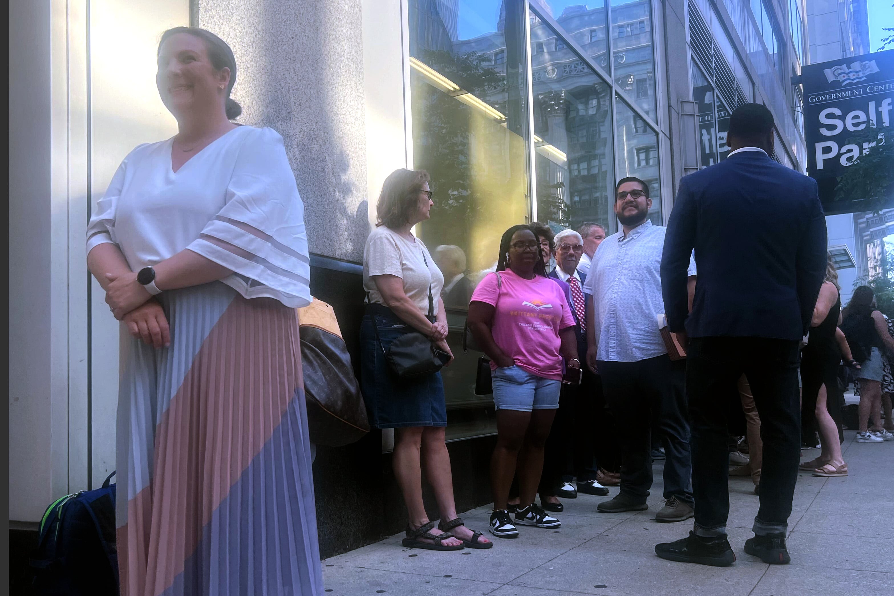 A woman in a white top stands in front of a line of people outside a building.