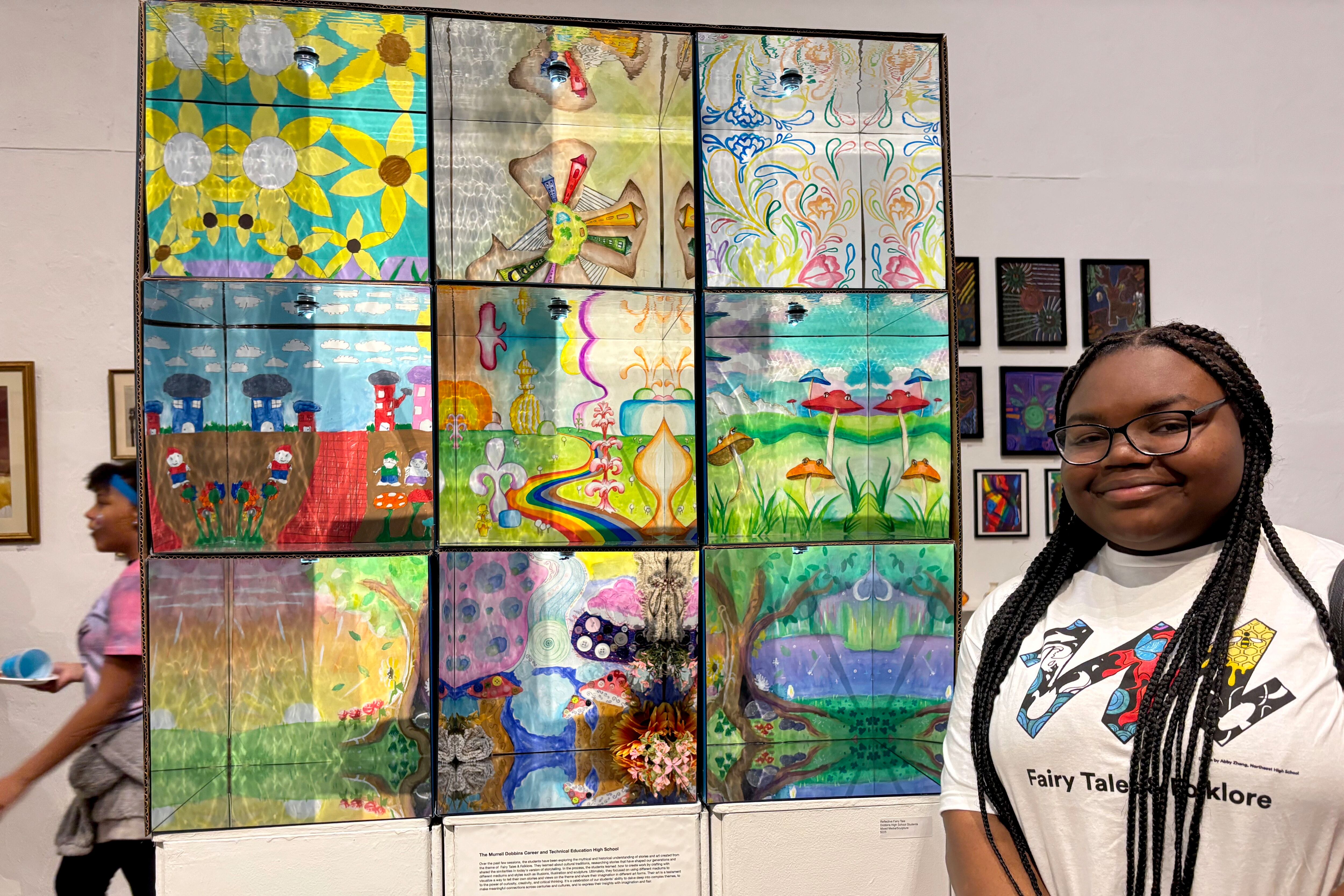 A high school senior with long dark braids and wearing a while t-shirt stands next to a piece of art while posing for a photograph.