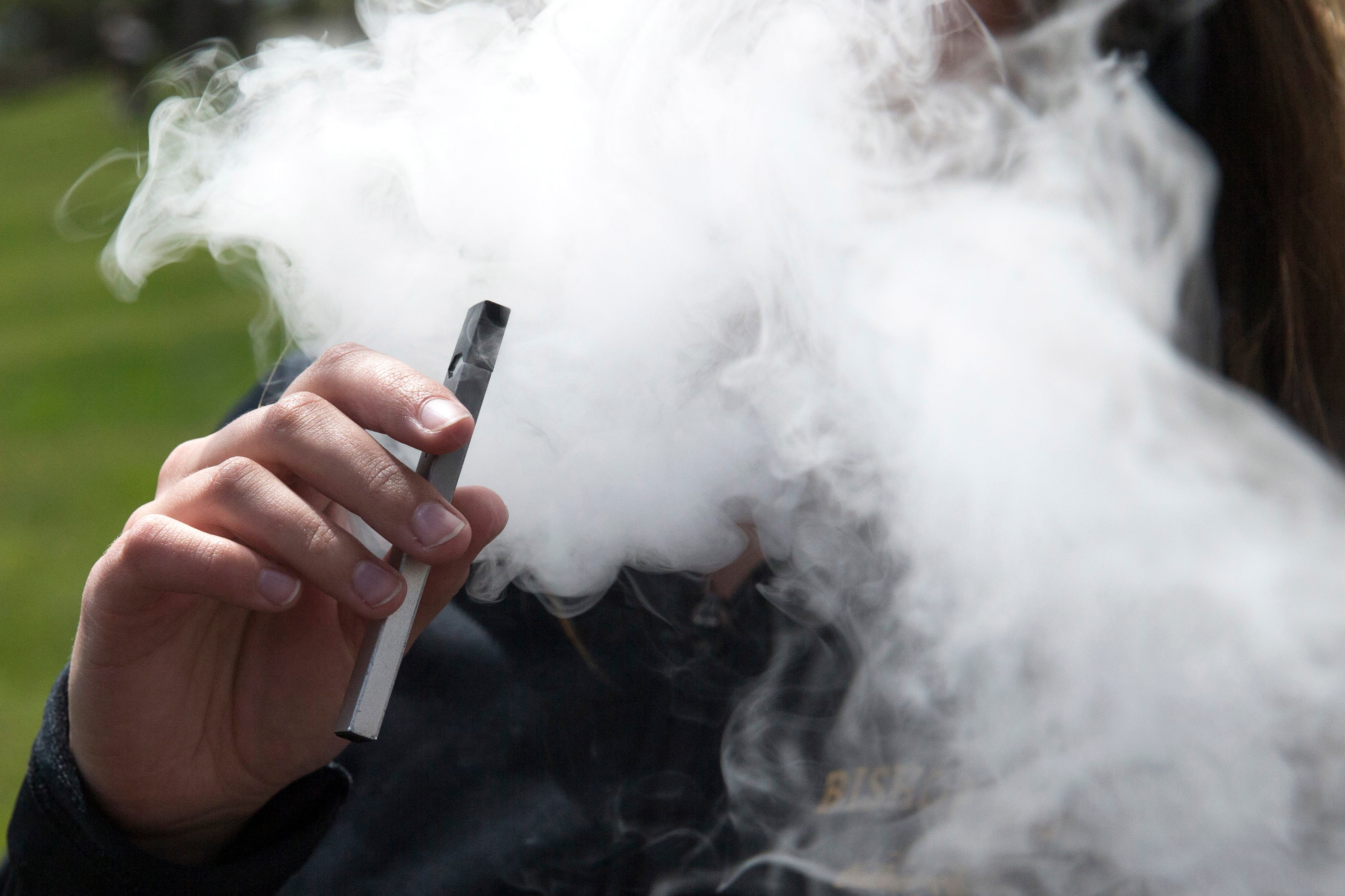 A teen holds an e-cigarette while a cloud of vape smoke covers their face outside.