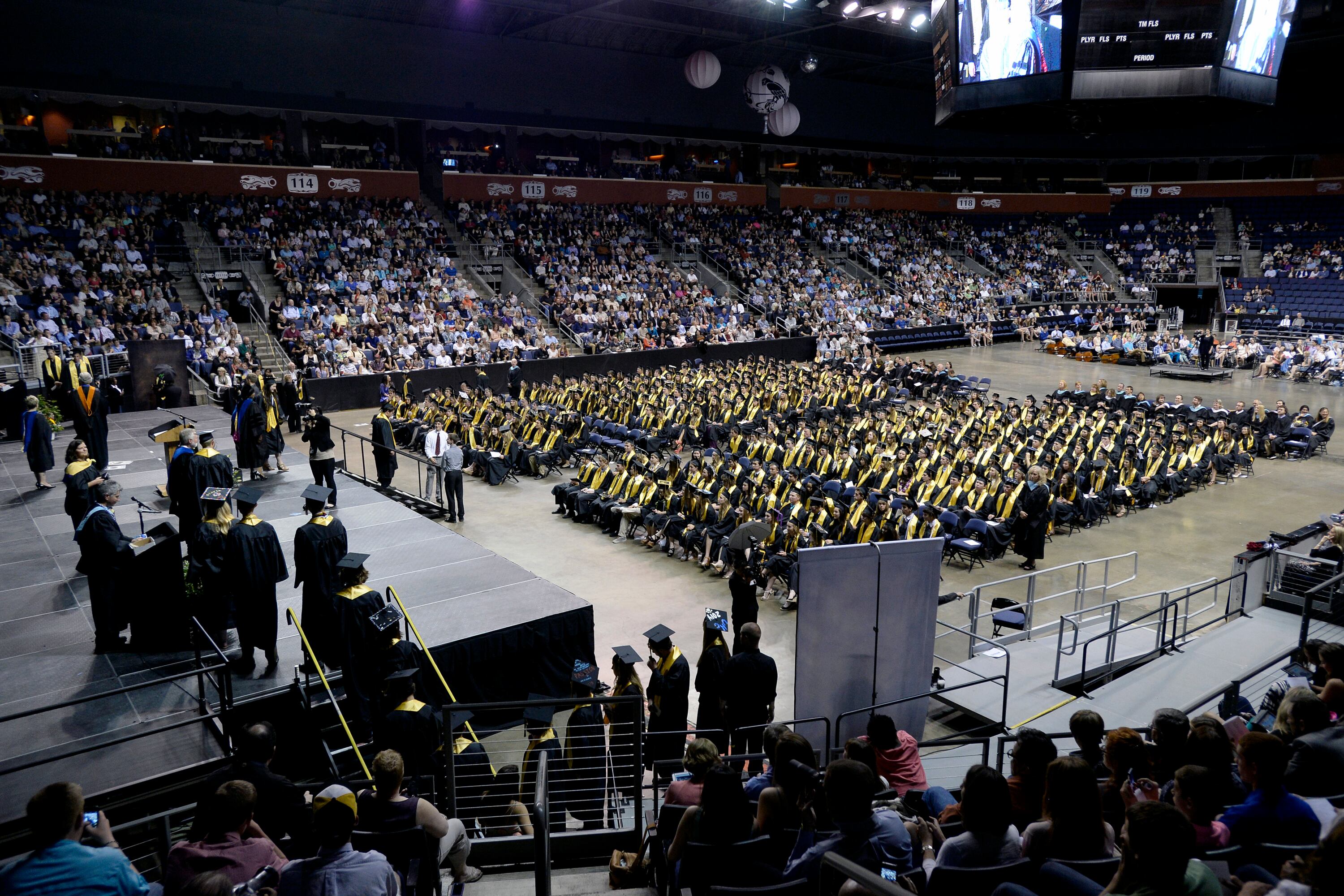 A large indoor stadium is full of high school graduates sitting in rows of chairs with friends and families sitting on the side.