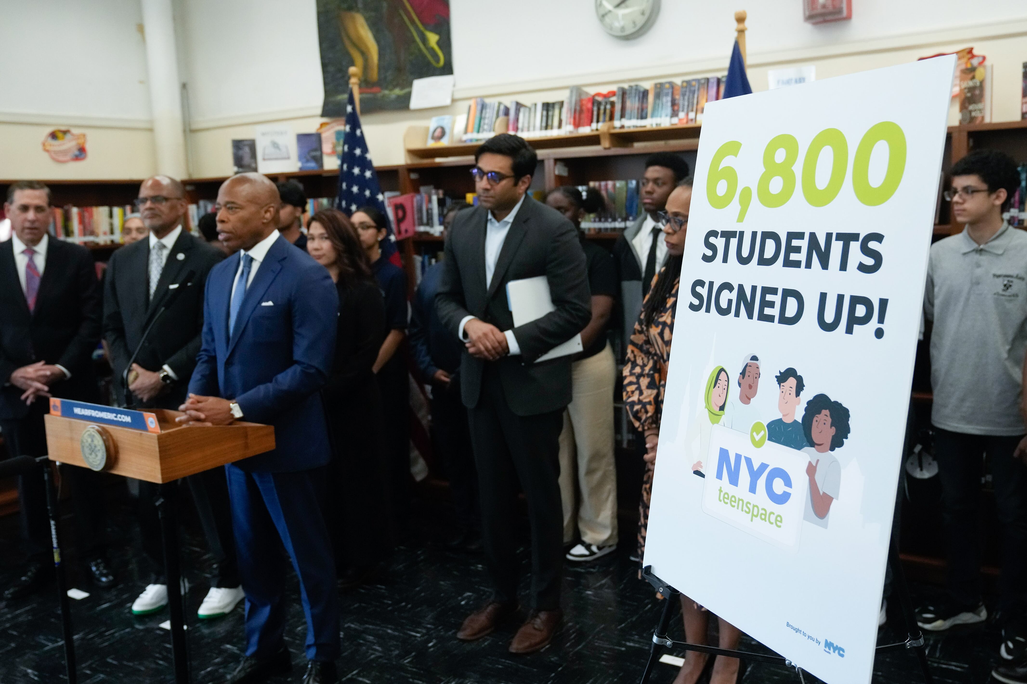 A group of people stand in front of a large bookshelf and behind a small wooden podium. there's a large white poster board with the words that say "6,800 students signed up."