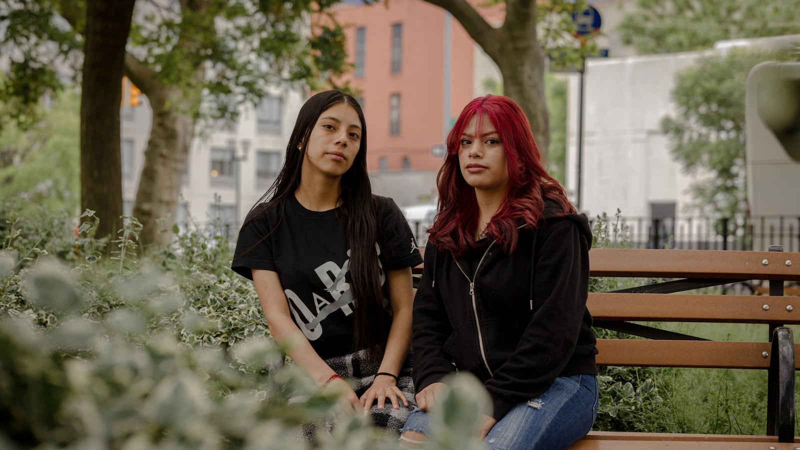 Two high school students and sisters, one with long dark hair and one with short red hair sit next to each other on a park bench with green trees and bushes around then.