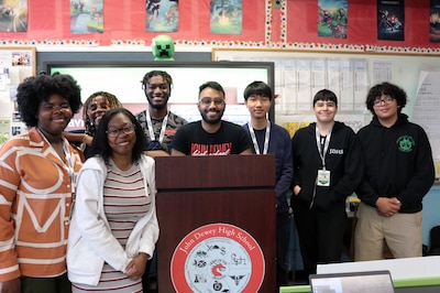 Des élèves et un enseignant posent pour une photo devant un tableau effaçable à sec et derrière un podium en bois dans une salle de classe.
