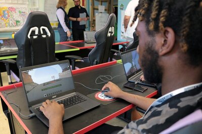 Un lycéen aux cheveux noirs travaille sur un ordinateur portable dans une salle de classe avec deux adultes en arrière-plan.