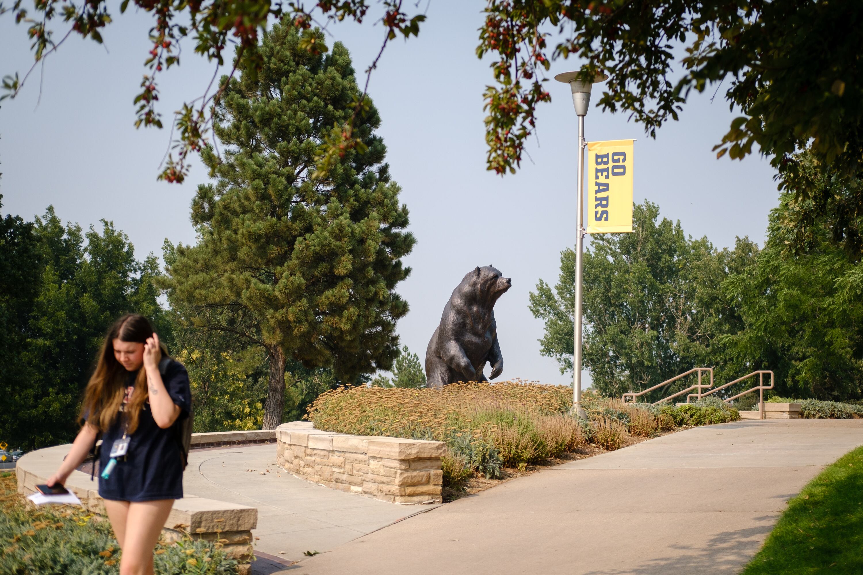 A student wearing a t-shirt and shorts walks down a sidewalk with a statue of a bear and green trees in the background.