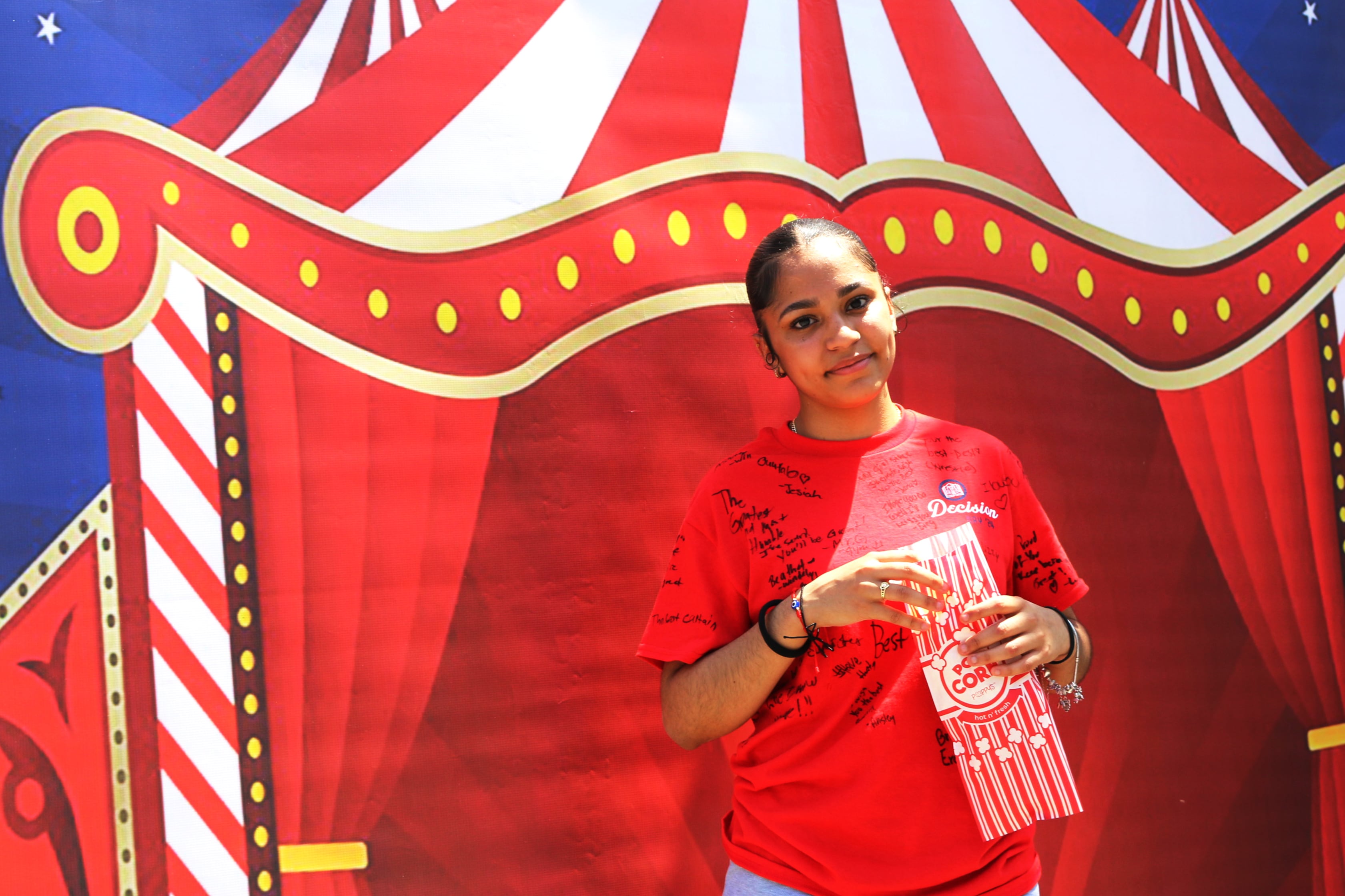 A girl wearing red holds popcorn in front of a circus-themed backdrop.