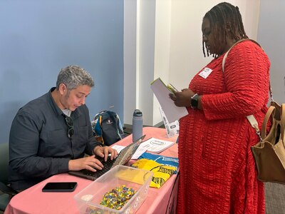 A man with short grey hair and wearing a dark dress shirt sits at a table while a woman with short dark hair and wearing a bright red dress stands on the other side of the table reading a pamphlet.