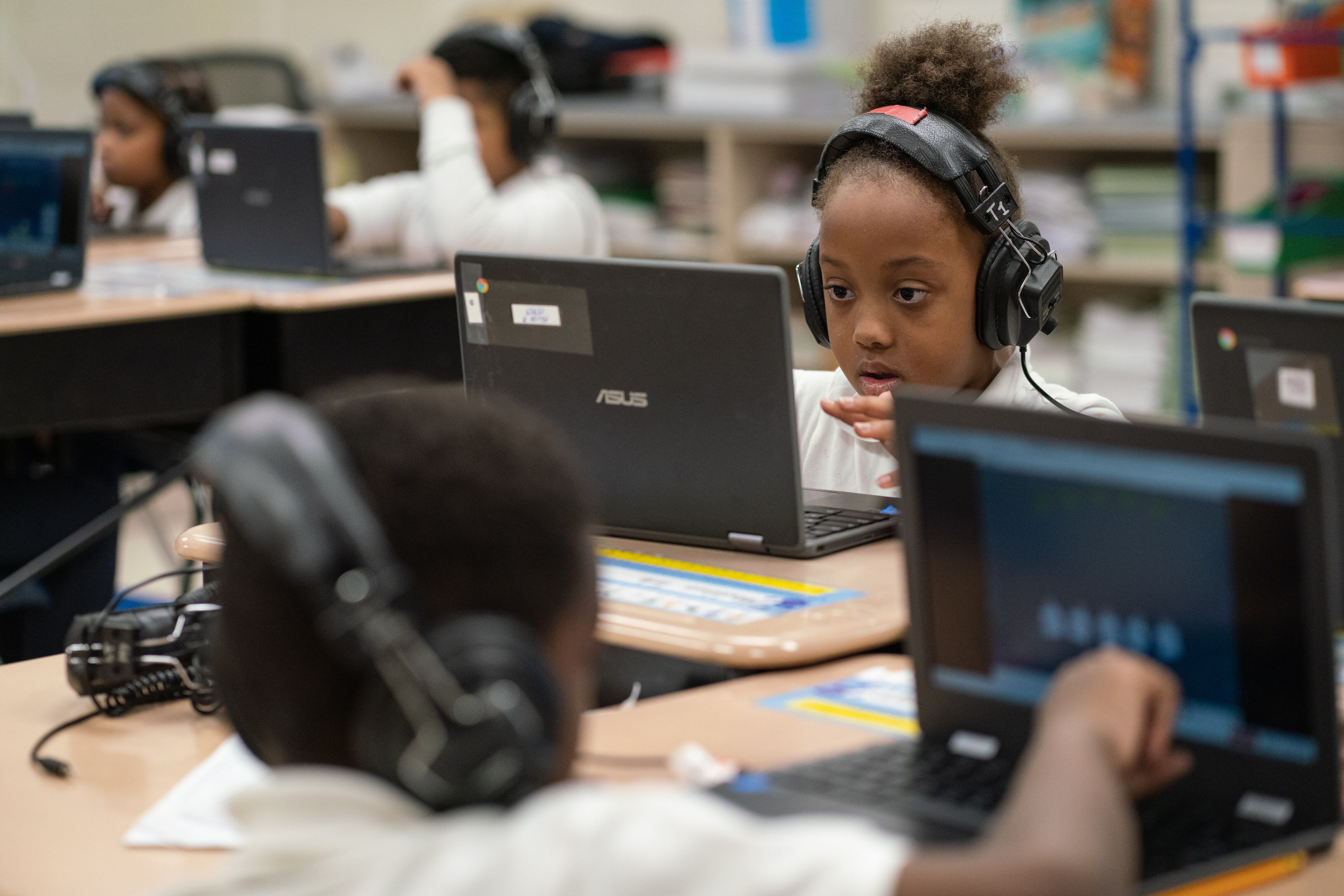 A student wearing headphones works at a laptop, surrounded by other students working in the classroom.