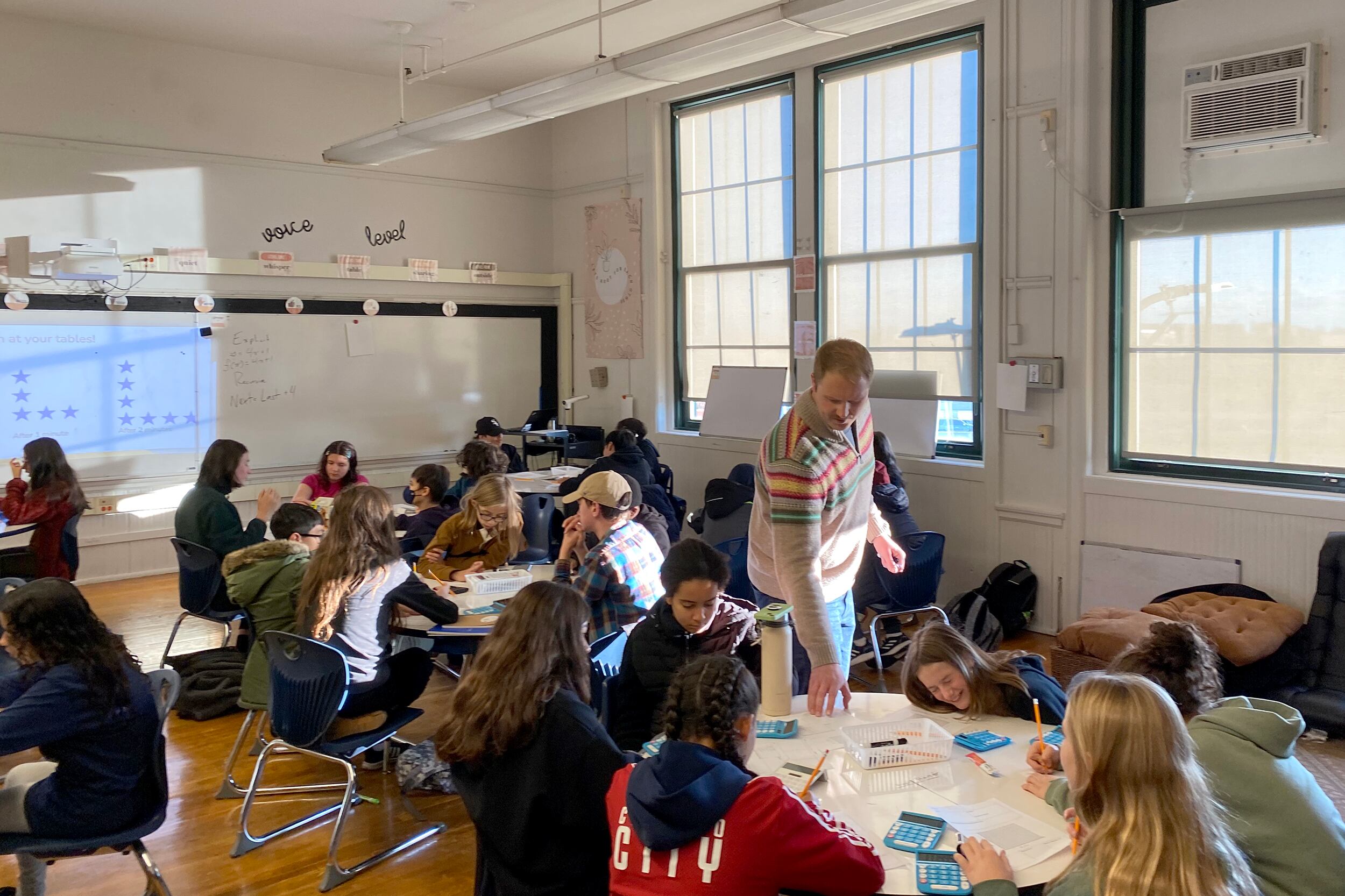 An adult teacher stands while a large group of students sit at their tables in a classroom with white walls in the background.