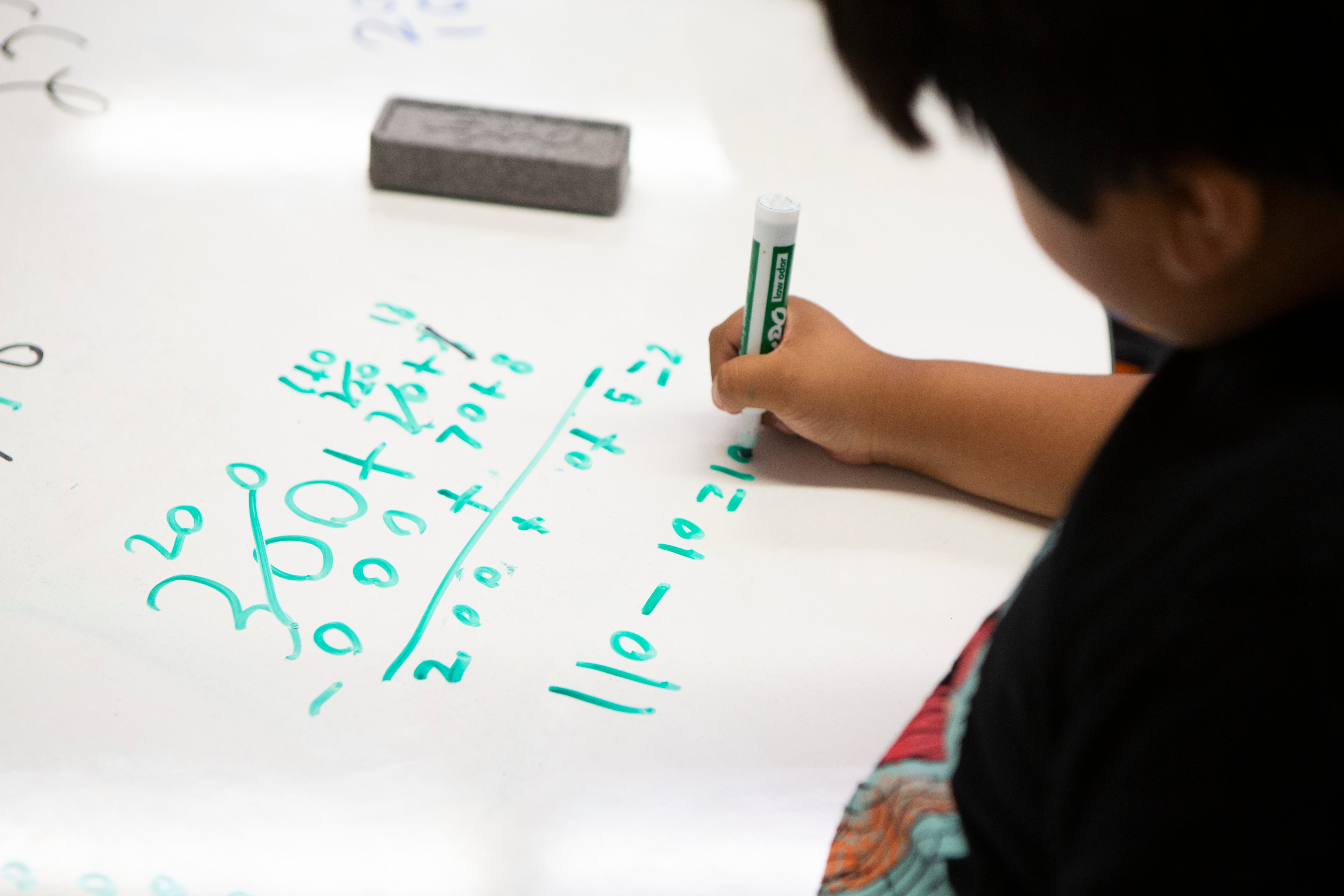 A young student writes numbers on a white board in green dry erase marker.