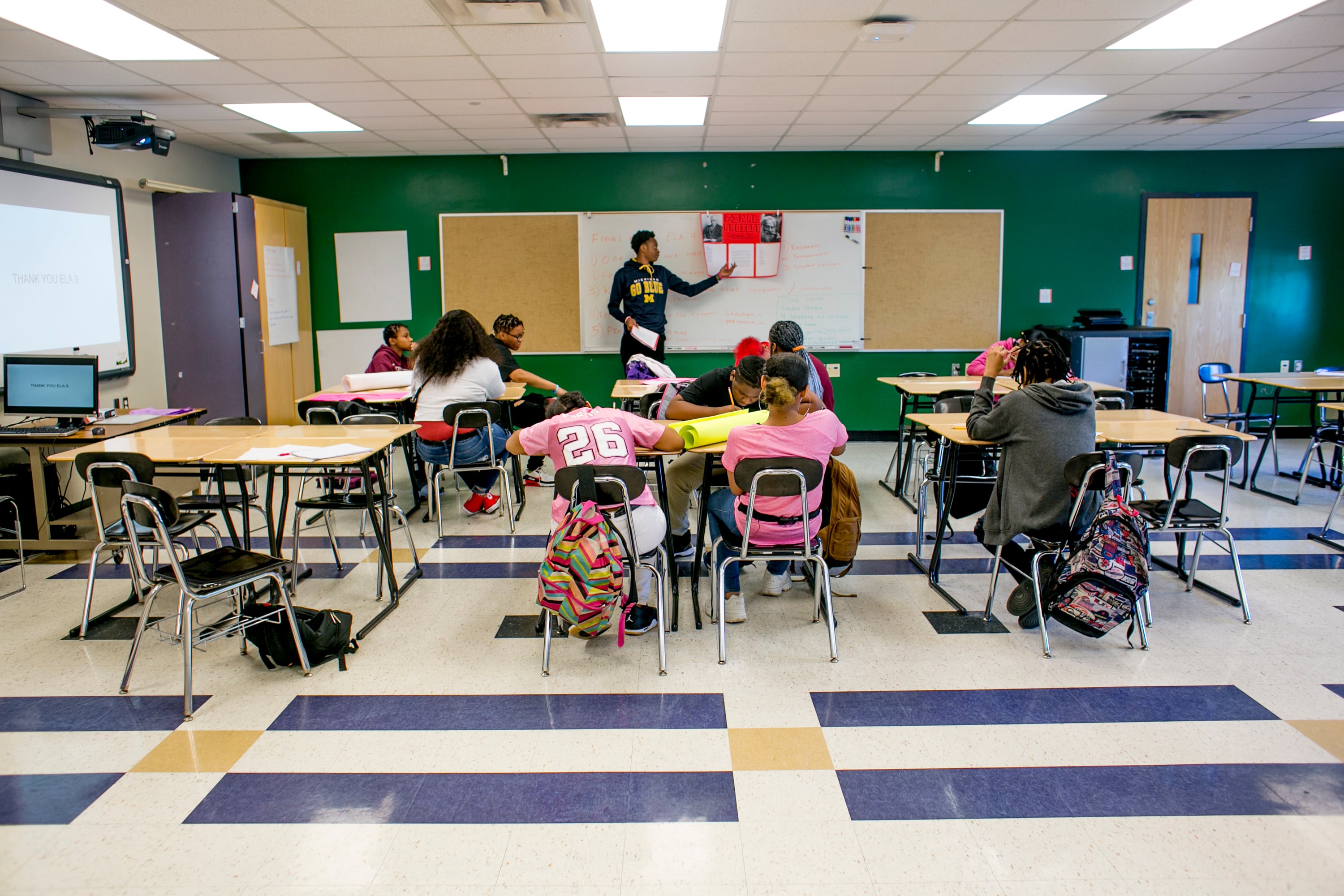 A student stands up in front of a small class of students sitting at wooden desks.