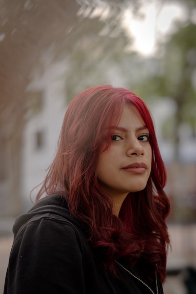 A high school senior with medium length red hair and wearing a black sweater poses for a portrait with green trees and a building in the background.