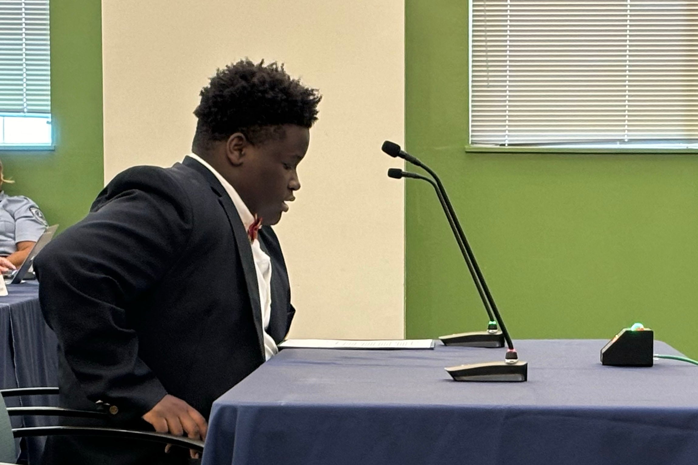 A high school student with short dark hair and wearing a dark suit sits at a table while speaking into a microphone with a white and green wall in the background.