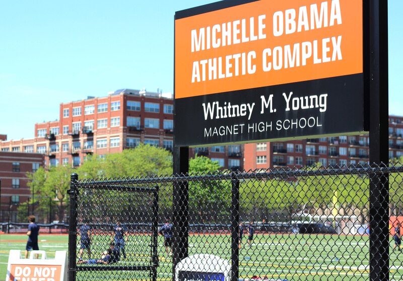 A large orange sign with white words stands in front of an athletic field outside.