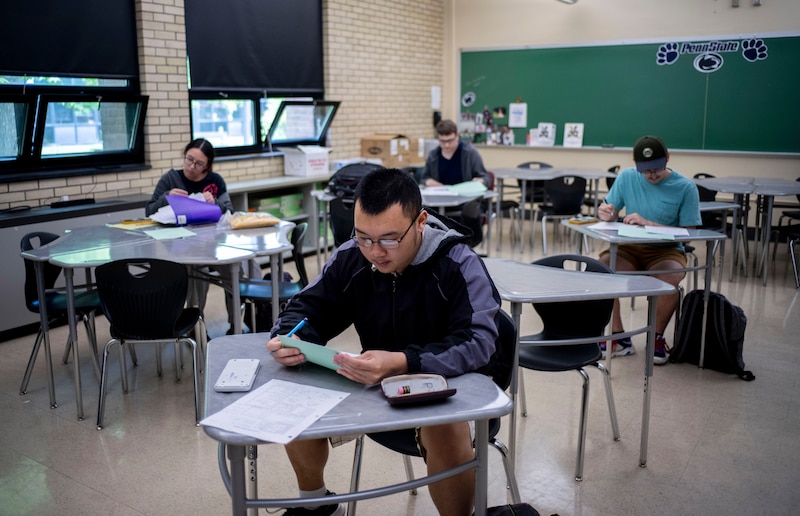 A student in glasses and a sweatshirt sits at a table in a classroom.