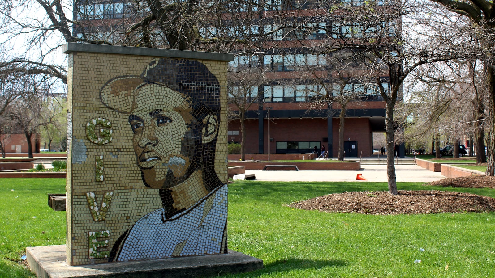 A large stone collage portrait of a male baseball player sits in a green lawn with a large building in the background.