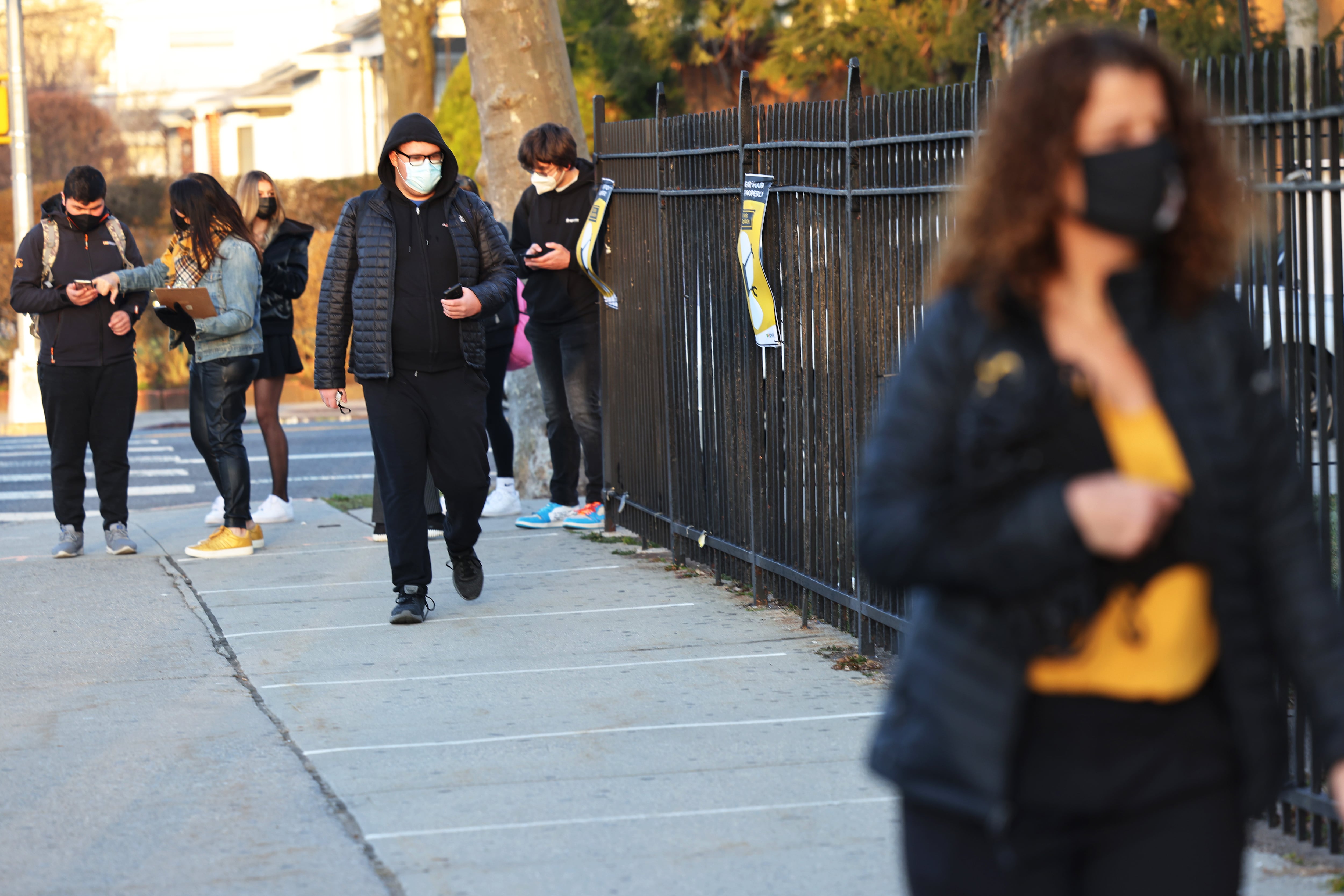 Teens wearing puffy coats and masks walk by a fence in front of a school.