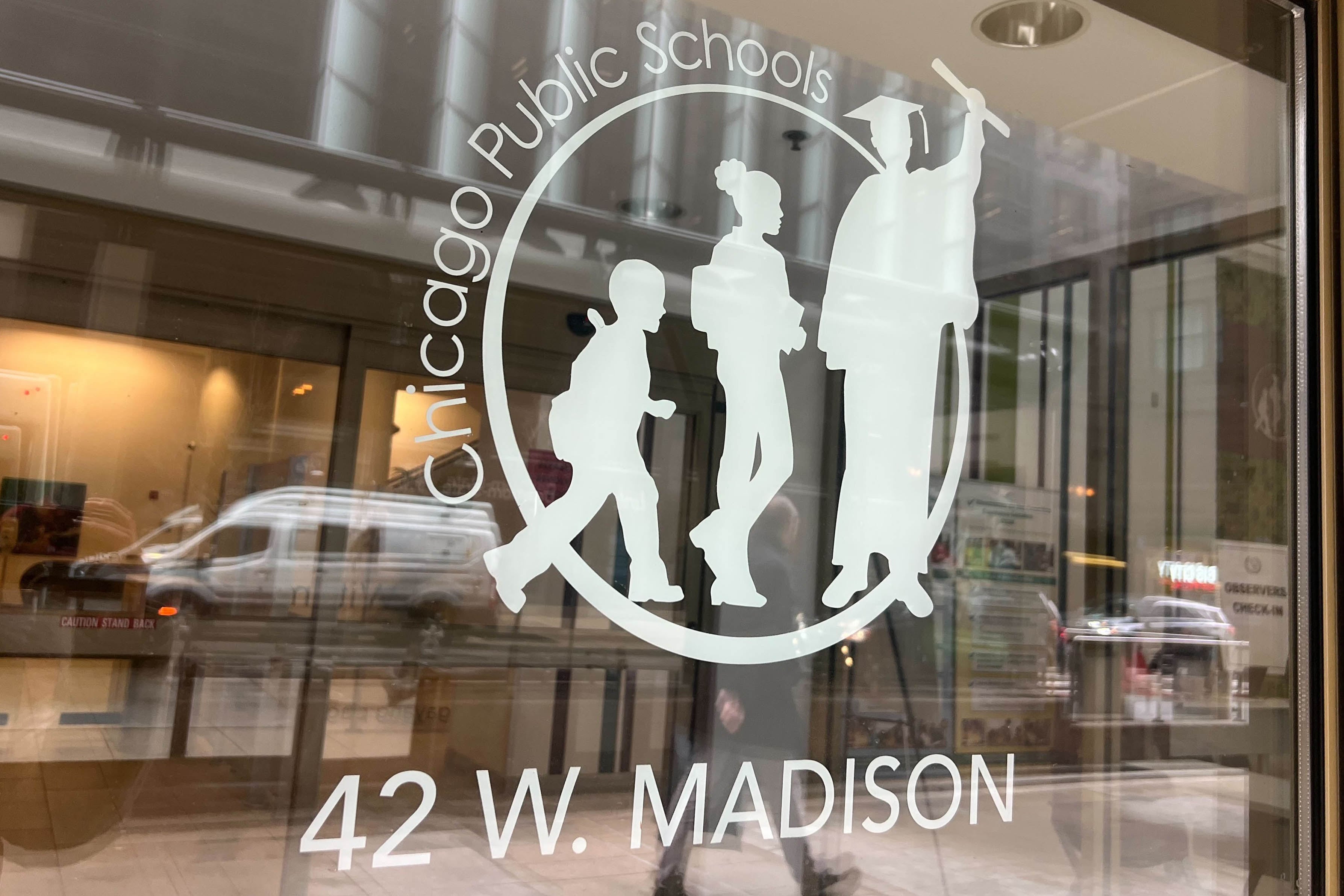 The Chicago Public Schools logo on the side of a glass wall with reflection of cars and people passing in the background.