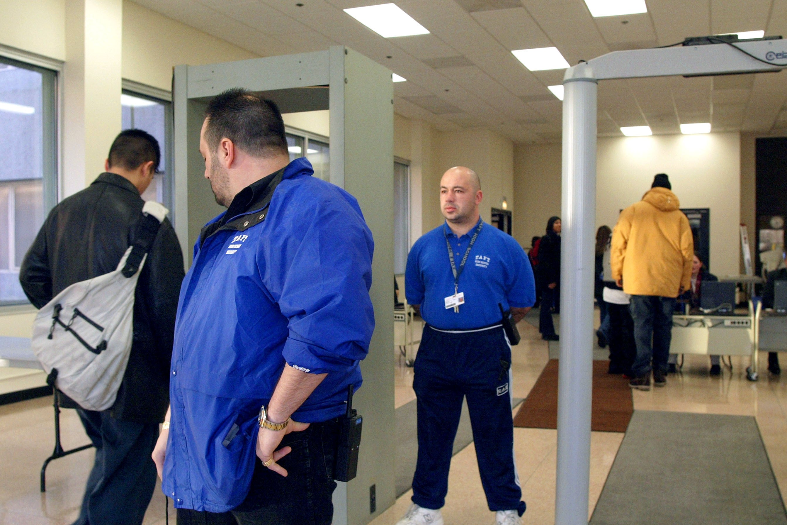 Two men in blue jackets stand in between two metal detectors while two high school students walk through.