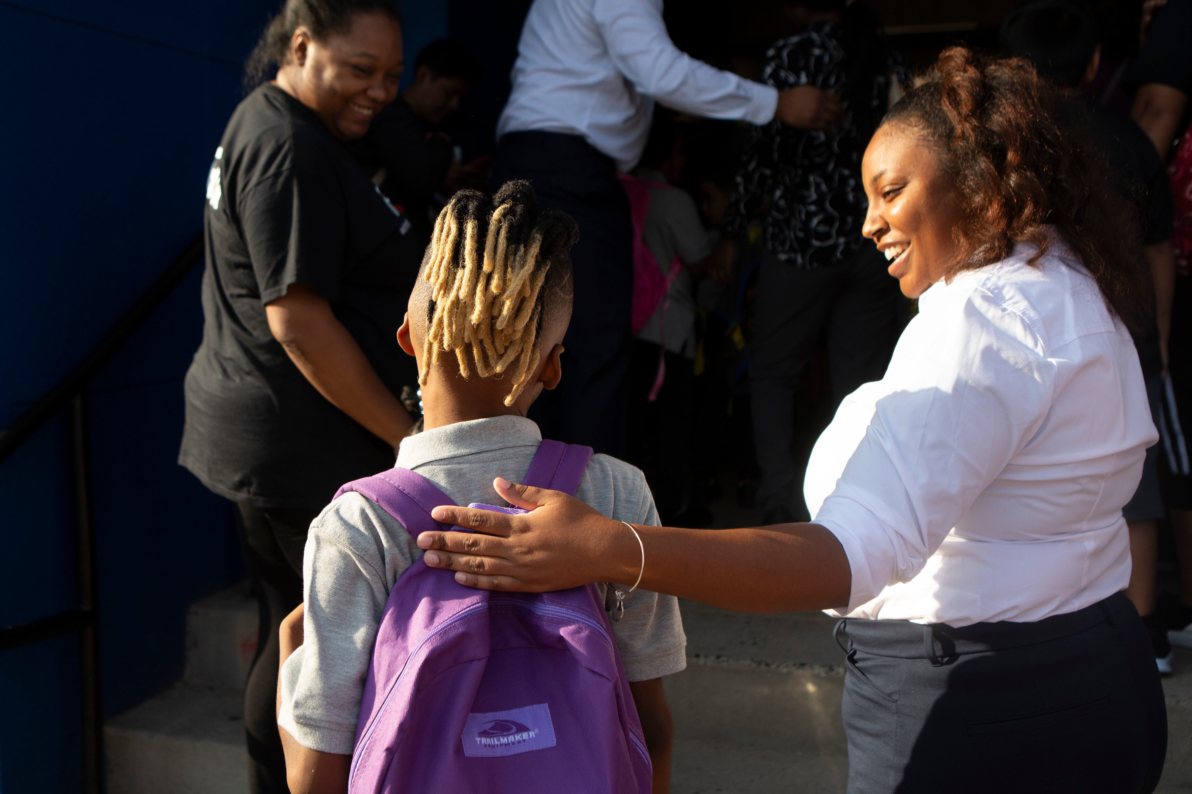 An adult with long hair and wearing a white blouse places her hand on a student's shoulder. The student has long hair in a ponytail and wearing a purple backpack.